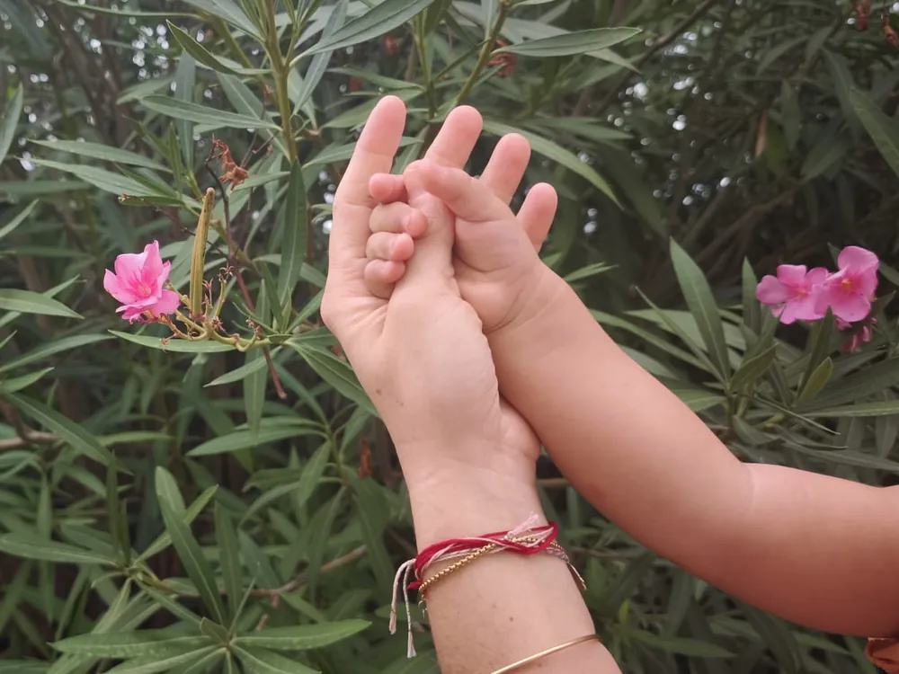 Main d'un parent tenant la main de son bébé en extérieur, devant un laurier-rose en fleurs